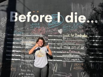 Smiling person giving thumbs-up in front of a large outdoor "Before I die..." interactive chalkboard wall filled with colorful handwritten wishes and messages.