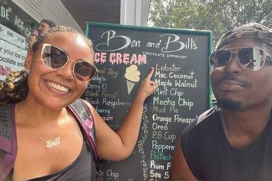 Two people in sunglasses smiling and pointing at an outdoor ice cream shop chalkboard menu with playful handwritten flavors like lobster, macadamia coconut and mint chip.