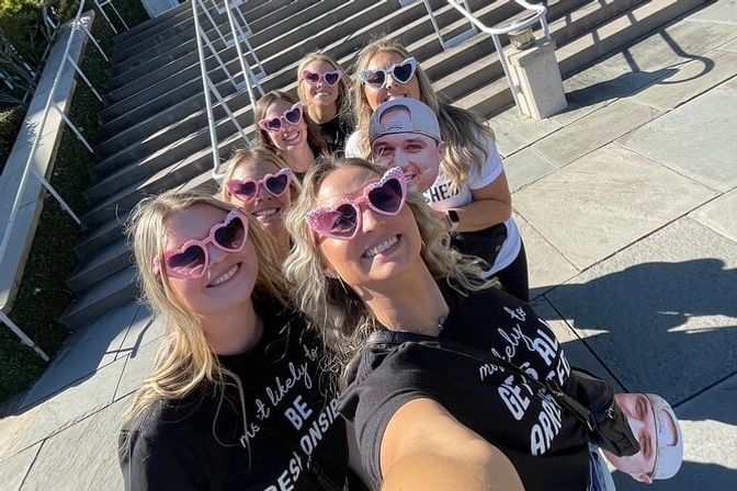 Group selfie of smiling women wearing pink heart-shaped sunglasses and matching shirts on sunlit stone steps outdoors