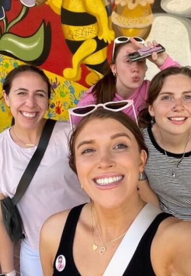 Four friends taking a cheerful selfie in front of a colorful handprint-and-bee street-art mural outdoors