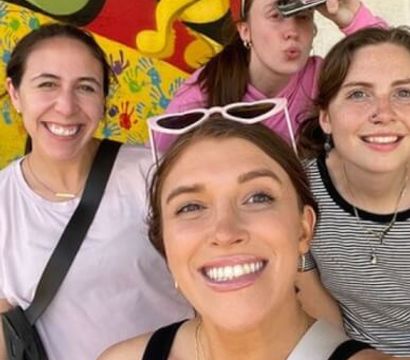 Four friends taking a cheerful selfie in front of a colorful handprint-and-bee street-art mural outdoors