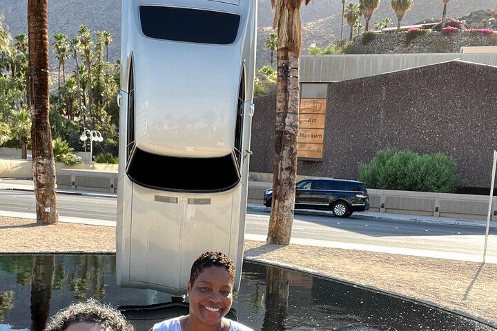 Two smiling visitors pose in front of a vertical silver car art installation rising from a reflecting pool, with palm trees, desert mountains and a museum building in the sunny plaza.