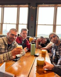 Four friends sharing hazy craft beers around a wooden communal table in a bright mountain-view brewery tasting room with large windows and string lights.