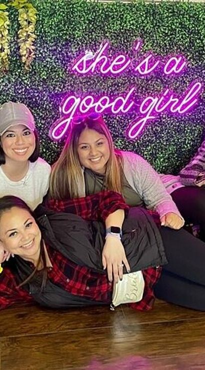 Group photo of six smiling women posing at an indoor leafy floral backdrop with a pink neon sign reading “she’s a good girl”; one woman lounges in front, casual outfits and sneakers.
