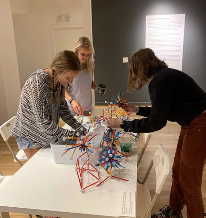 Three visitors at an indoor museum gallery table building colorful stick-and-ball geometric sculptures — hands-on interactive STEM exhibit with spiky red, blue and yellow models and a wall text panel in the background.