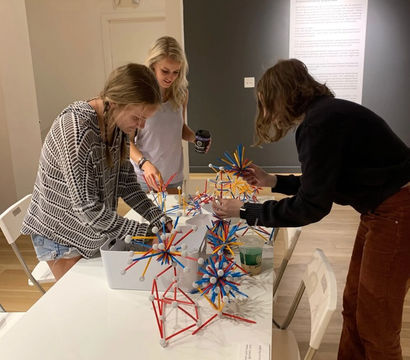 Three visitors at an indoor museum gallery table building colorful stick-and-ball geometric sculptures — hands-on interactive STEM exhibit with spiky red, blue and yellow models and a wall text panel in the background.