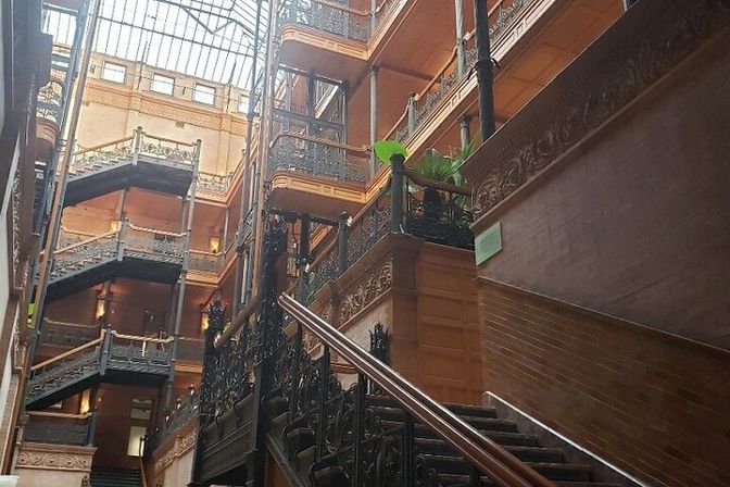 Grand historic atrium featuring ornate wrought-iron railings, a sweeping dark-wood staircase, stacked balconies and a soaring glass skylight overhead.