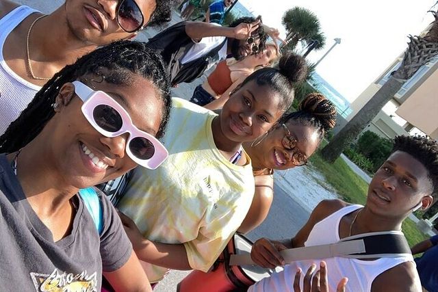 Smiling group selfie of six friends on a sunny beachside street with palm trees and ocean in the background, wearing sunglasses and casual summer clothes
