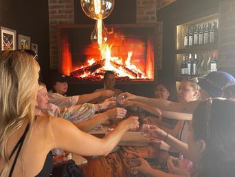 Group of friends toasting with small glasses around a rustic wooden table in a cozy wine bar, glowing fireplace, exposed brick and shelves of wine bottles