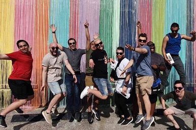 Group of men jumping and posing in front of a rainbow-colored yarn wall mural on a sunny urban street, vibrant street art group photo.