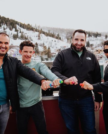 Four friends on a rooftop balcony in a snowy mountain town, smiling and bumping fists decorated with colorful candy ring toys with snow-covered hills and houses in the background.