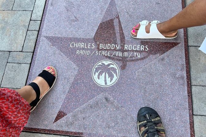 Red granite sidewalk star plaque honoring an entertainer (Radio/Stage/Film/TV) with a palm-tree emblem, surrounded by three people’s sandal-clad feet on tiled pavement.