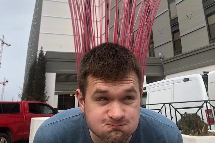 Playful portrait of a young man in a blue shirt puffing his cheeks outdoors in front of a tall red metal sculpture and a modern building entrance with parked vans and a red pickup