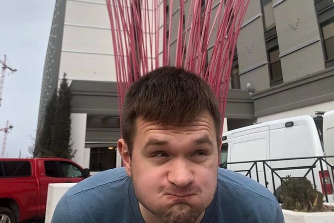 Playful portrait of a young man in a blue shirt puffing his cheeks outdoors in front of a tall red metal sculpture and a modern building entrance with parked vans and a red pickup