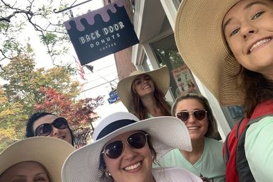 Cheerful group selfie of five friends in wide-brim sun hats and sunglasses smiling outside a donut shop on a tree-lined main street with autumn foliage.