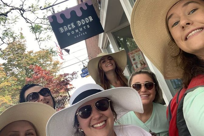 Cheerful group selfie of five friends in wide-brim sun hats and sunglasses smiling outside a donut shop on a tree-lined main street with autumn foliage.