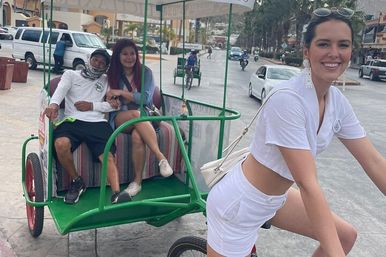 Smiling woman in white pedals a green pedicab along a sunny coastal promenade with two relaxed passengers in the back.