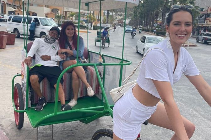 Smiling woman in white pedals a green pedicab along a sunny coastal promenade with two relaxed passengers in the back.