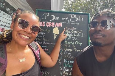 Two smiling people in sunglasses pose and point at a colorful handwritten chalkboard listing ice cream flavors outside a sunny shop