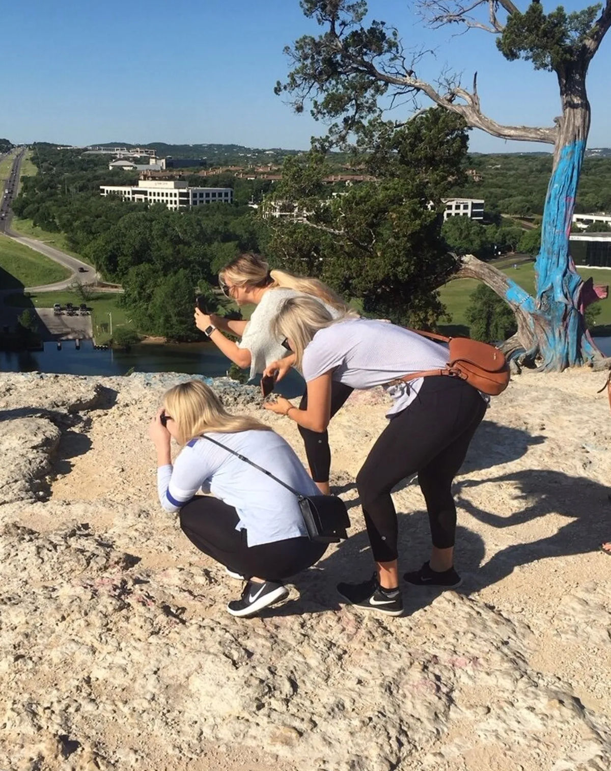 Three women crouch and lean to snap photos from a rocky cliffside overlook above a river and tree-lined valley, with a painted tree trunk and distant low-rise buildings under a clear blue sky.