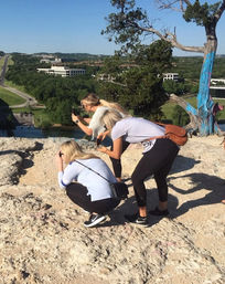 Three women crouch and lean to snap photos from a rocky cliffside overlook above a river and tree-lined valley, with a painted tree trunk and distant low-rise buildings under a clear blue sky.