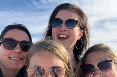 Four smiling women in sunglasses taking a close-up outdoor selfie on a sunny day with blue sky and sandy beach reflected in their lenses