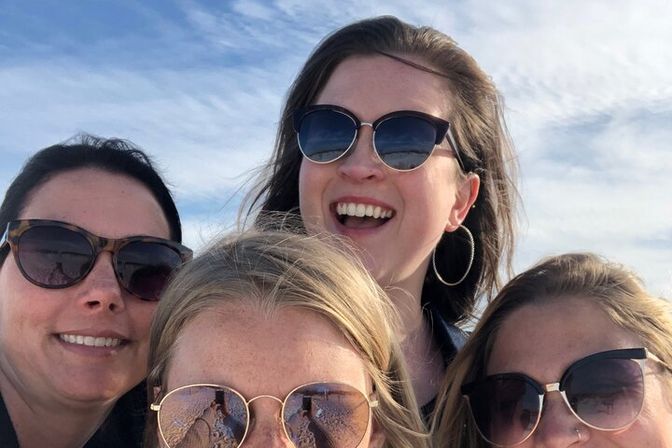 Four smiling women in sunglasses taking a close-up outdoor selfie on a sunny day with blue sky and sandy beach reflected in their lenses