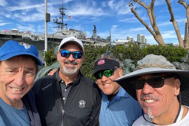 Four smiling men in hats and sunglasses take a selfie at the San Diego waterfront park with a docked aircraft carrier, bronze sculptures and bright blue sky