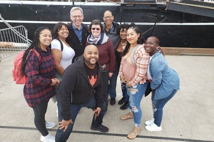 Smiling diverse group of adults posing for a casual group photo on a concrete pier in front of a black-hulled boat at a waterfront