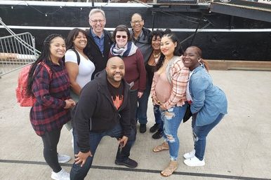 Smiling diverse group of adults posing for a casual group photo on a concrete pier in front of a black-hulled boat at a waterfront
