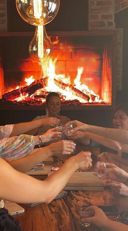 Group of people seated around a rustic wooden table raising shot glasses in a cheerful toast, cozy bar interior with a glowing fireplace and wine bottles on shelves.