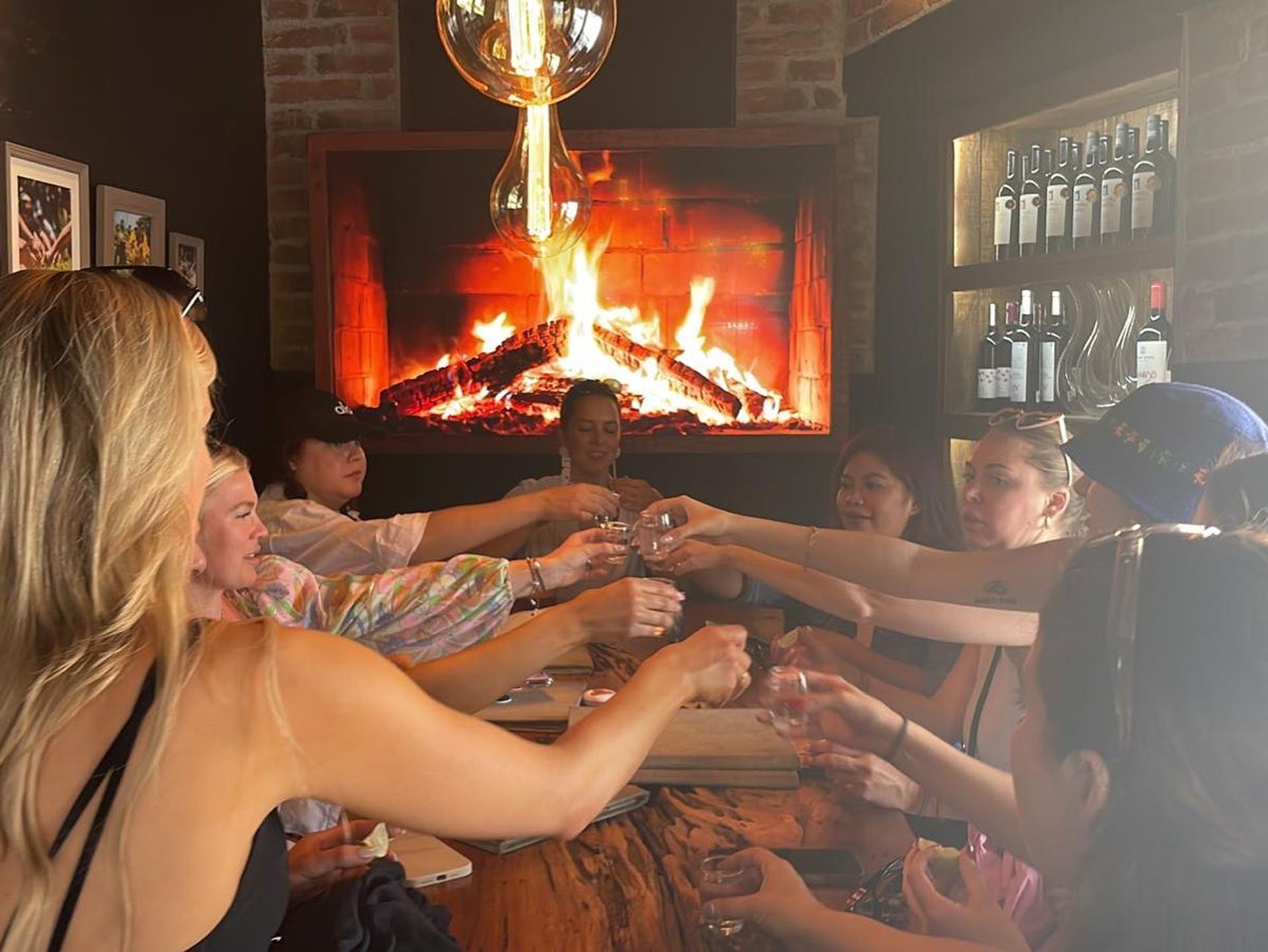 Group of people seated around a rustic wooden table raising shot glasses in a cheerful toast, cozy bar interior with a glowing fireplace and wine bottles on shelves.