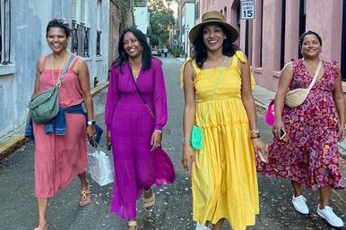 Four smiling women in colorful summer dresses (coral, magenta, yellow with straw hat, floral) walking together on a narrow pastel-painted urban street for a sunny stroll.