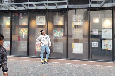 Person wearing a HOPE sweatshirt and bright sneakers poses on a city sidewalk in front of a downtown storefront window displaying framed artwork and award ribbons.