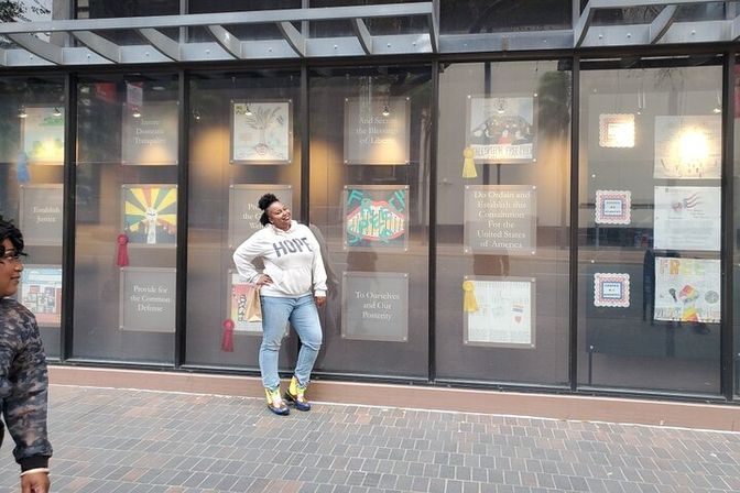 Person wearing a HOPE sweatshirt and bright sneakers poses on a city sidewalk in front of a downtown storefront window displaying framed artwork and award ribbons.