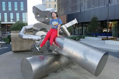 Smiling person in red leggings and blue T‑shirt sitting atop a large stainless‑steel robot sculpture in a downtown urban plaza with modern office buildings