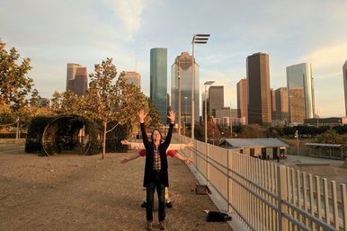 Three people playfully posing with layered outstretched arms on a park overlook framed by trees and the Houston downtown skyline at golden hour