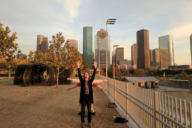 Three people playfully posing with layered outstretched arms on a park overlook framed by trees and the Houston downtown skyline at golden hour