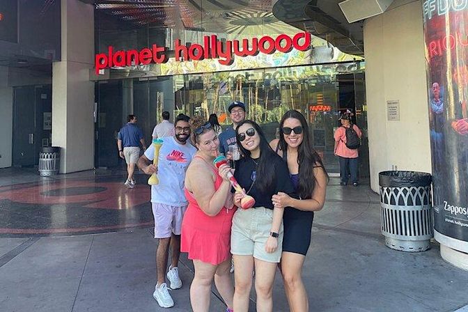 Group of five young adults smiling on an entertainment-district sidewalk in front of a glass entrance with a large red neon sign, wearing sunglasses and holding colorful souvenir yard cups — daytime tourist scene.