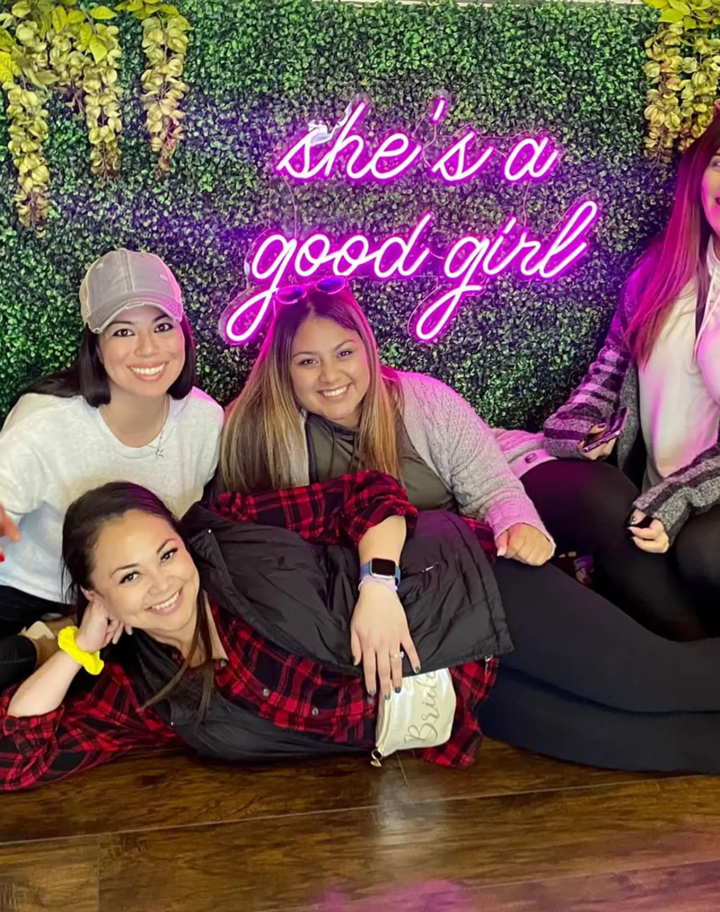 Group of smiling friends posing on the floor in front of a faux boxwood wall with a neon pink sign that reads "she's a good girl" — playful indoor photo spot with casual outfits.