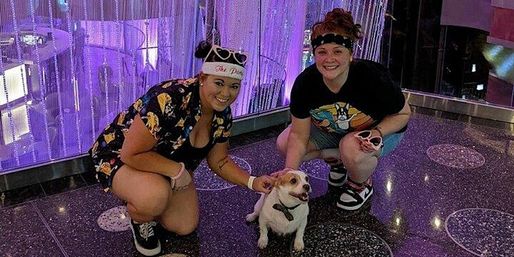 Two people crouch and smile while petting a small white-and-tan dog in a purple LED-lit indoor entertainment space with hanging bead curtains and a speckled floor.