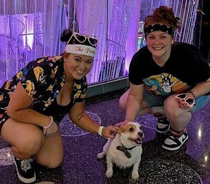 Two people crouch and smile while petting a small white-and-tan dog in a purple LED-lit indoor entertainment space with hanging bead curtains and a speckled floor.