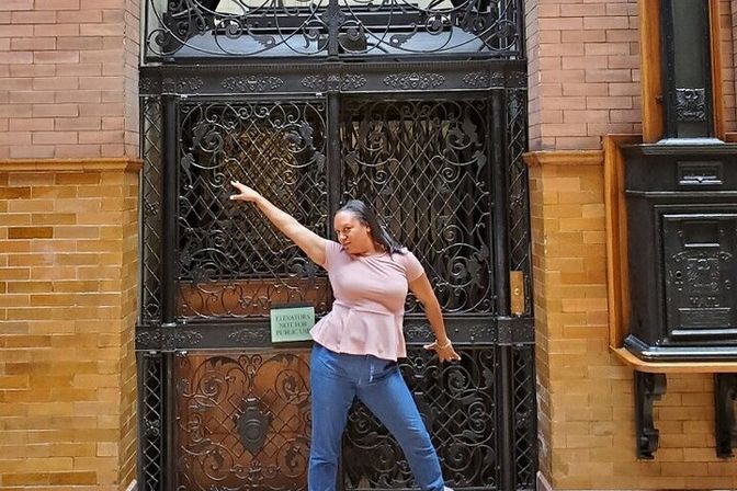 Woman in a pink peplum top and jeans striking a playful pose, pointing up in front of an ornate wrought-iron gate inside a historic brick building lobby.