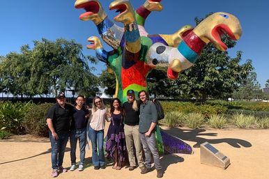 Six adults smiling and posing in front of a playful, colorful mosaic multi‑headed public art sculpture in a sunny outdoor park with trees and landscaping.