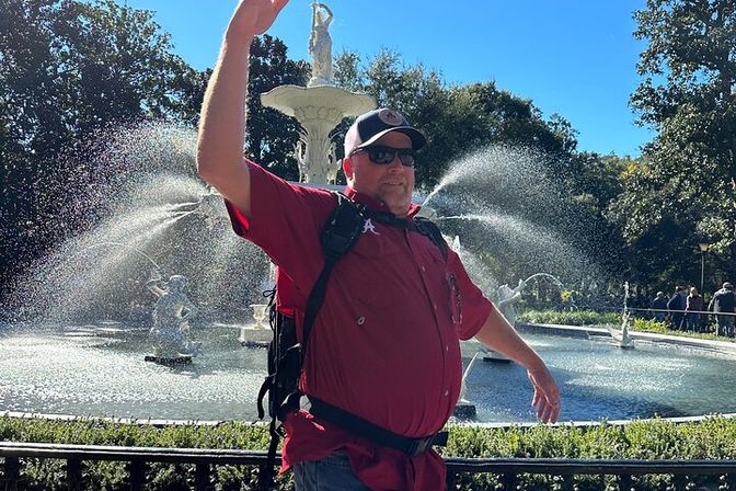 Person in a red shirt and baseball cap with a backpack, arms raised, posing in front of an ornate statue fountain spraying water in a sunny park setting.