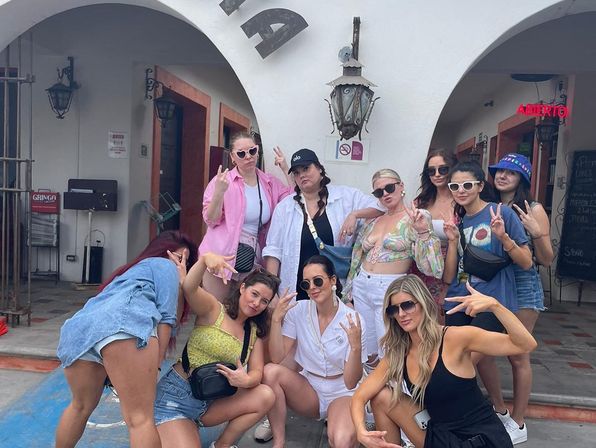 Group of friends striking playful poses and peace signs outside white stucco arches and hanging lanterns in a sunny beach-town plaza, wearing summer outfits and sunglasses.