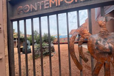 Rusted metal gate opening onto a contemporary outdoor sculpture garden with gravel desert landscaping, abstract bronze sculptures, drought-tolerant plants and a colorful mural on stucco walls under a partly cloudy sky.