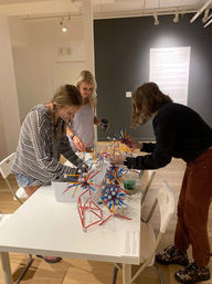 Three people at an indoor art gallery workshop building playful geometric sculptures from colorful red, blue, and yellow rods with white connectors on a white table; one person holds a coffee cup, hardwood floor and informational wall panel visible.