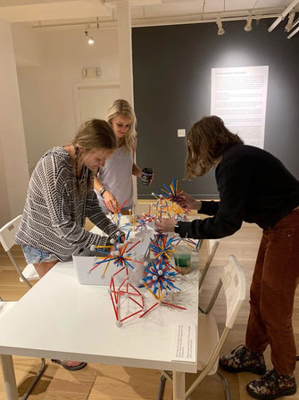 Three people at an indoor art gallery workshop building playful geometric sculptures from colorful red, blue, and yellow rods with white connectors on a white table; one person holds a coffee cup, hardwood floor and informational wall panel visible.
