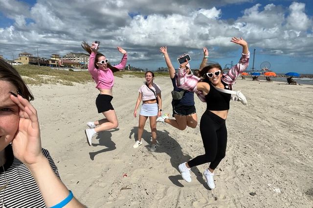 Group of friends jumping on a sandy beach under a partly cloudy blue sky, with a boardwalk and ferris wheel visible on the distant coastline — playful seaside vacation scene.
